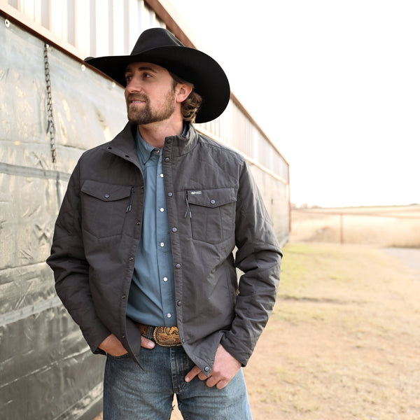 Man wearing a black jacket, blue shirt, and jeans standing against a wall.