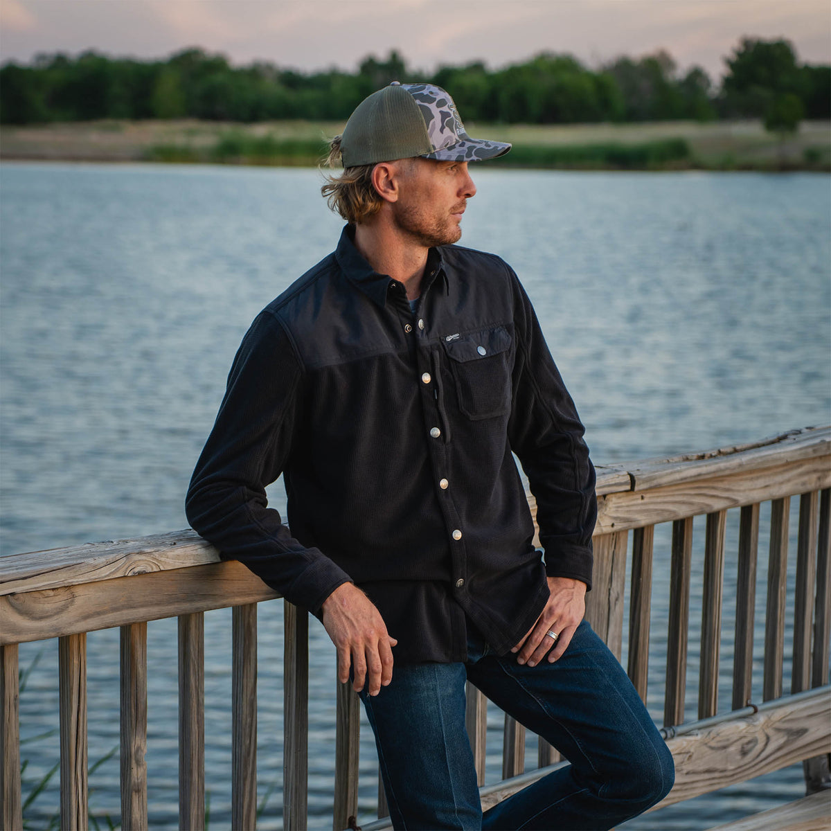 Man leaning on a wooden railing by a lake wearing a black button up shirt, jeans and a camo hat.