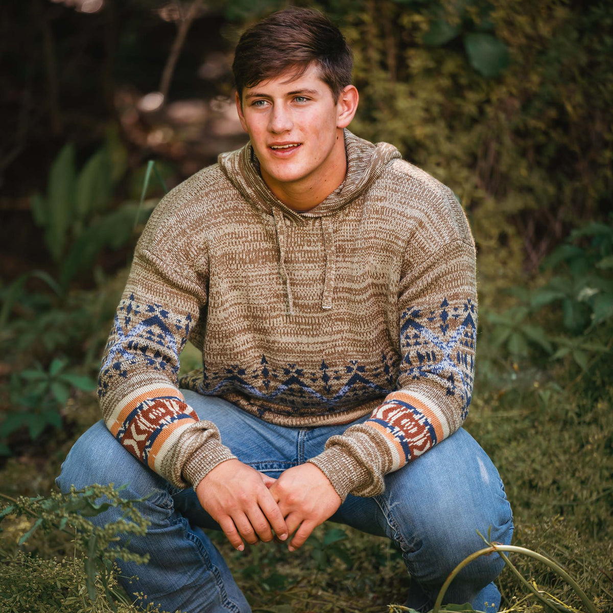 Man kneeling in the woods wearing blue jeans and a brown hoodie with a patterned design, drawstring hood and kangaroo pouch.