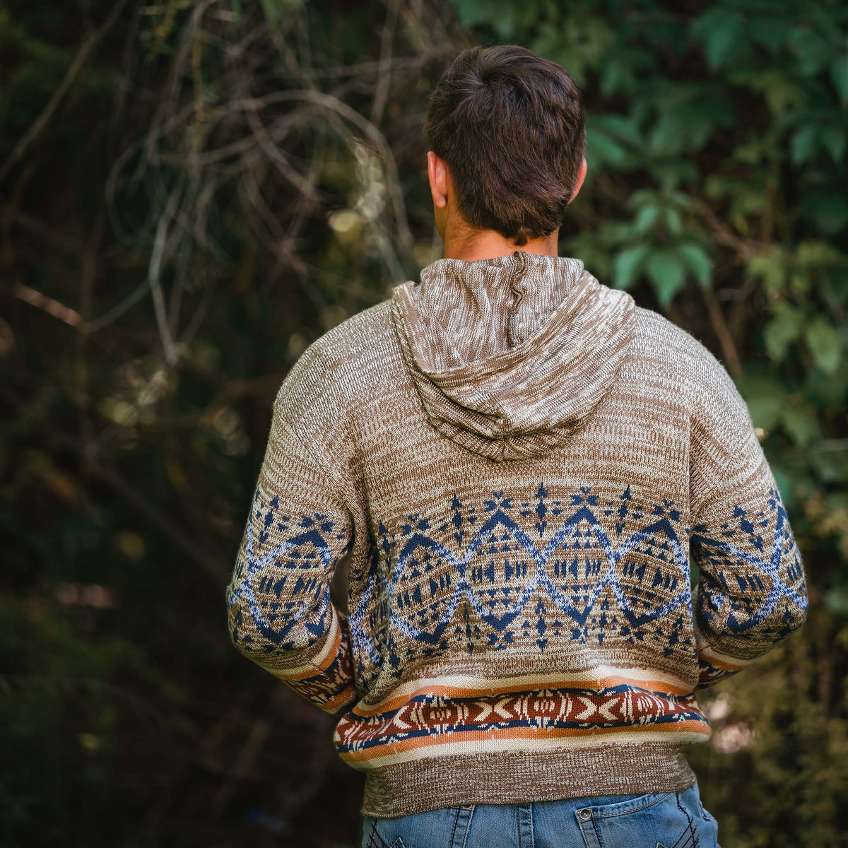 Person facing away wearing a brown hoodie with a patterned design and drawstring hood in the woods.