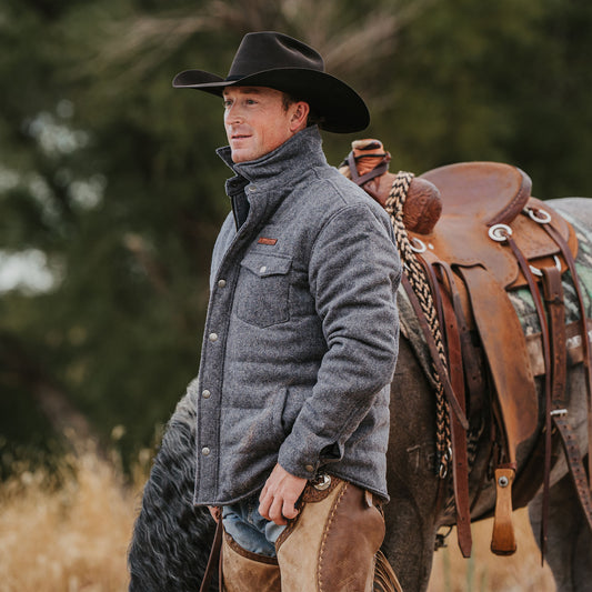 Man wearing a black cowboy hat and gray wool jacket standing in a field with a horse.