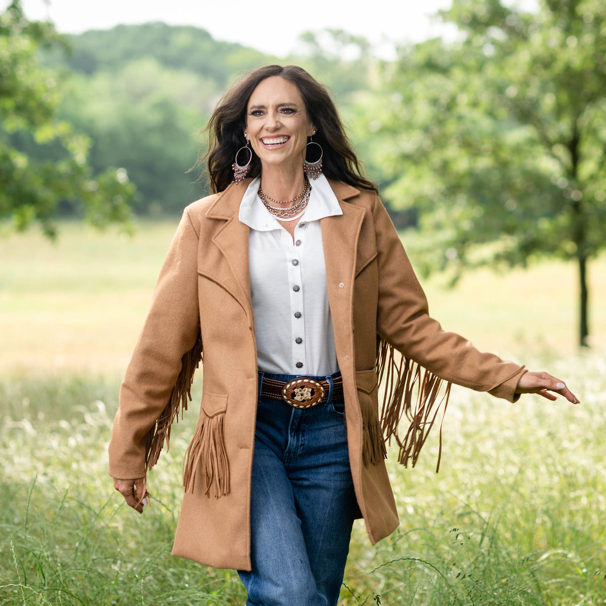 Jen Smith in an outdoor setting wearing jeans, a white collared shirt and a brown tailored wool overcoat for women with fringe accents on the pockets and sleeves.