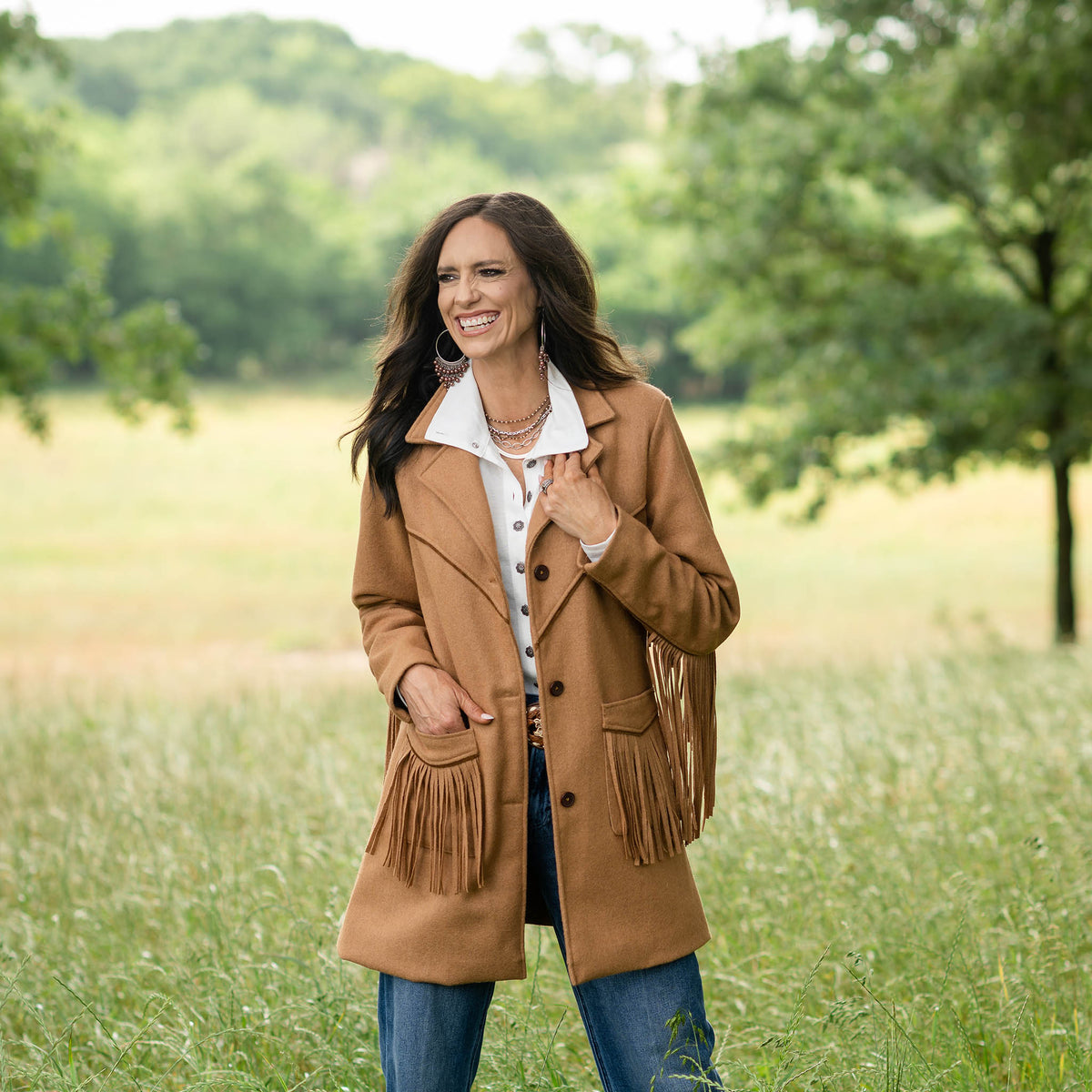 Jen Smith in an outdoor setting wearing jeans, a white collared shirt and a brown tailored wool overcoat for women with fringe accents on the pockets and sleeves.