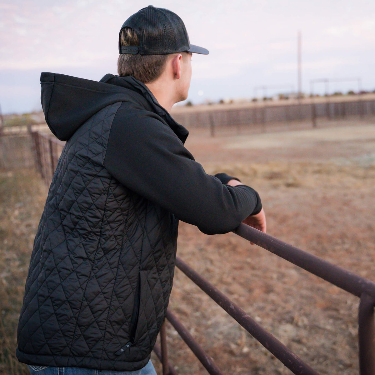 Man leaning against a metal fence on a ranch wearing a gray hat and black quilted jacket with a hood, jersey knit sleeves, and zip closure.
