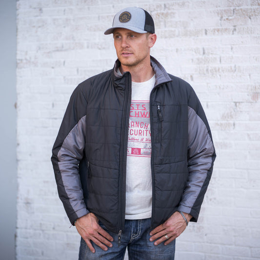 Man wearing a gray hat, jeans and a men's black and gray puffer jacket with a zip front closure in front of a brick wall.