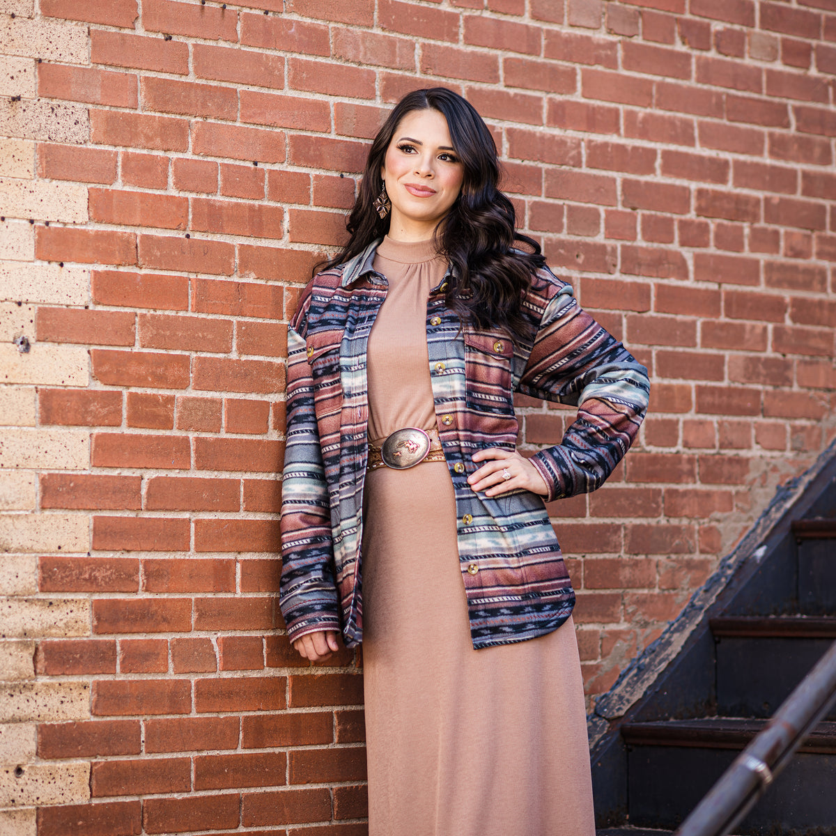 Woman wearing a tan dress and multicolored striped women's shirt jacket with button front closure and collar, featuring four exterior pockets leaning against a brick building.