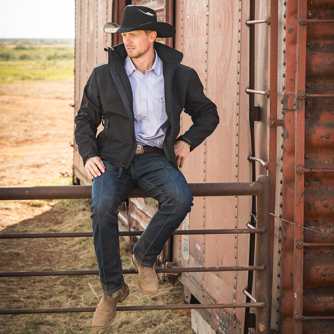 Man in cowboy attire sitting on a metal gate with a rustic background wearing a black jacket with front chest zip pocket, front zip closure and hand warming pockets 