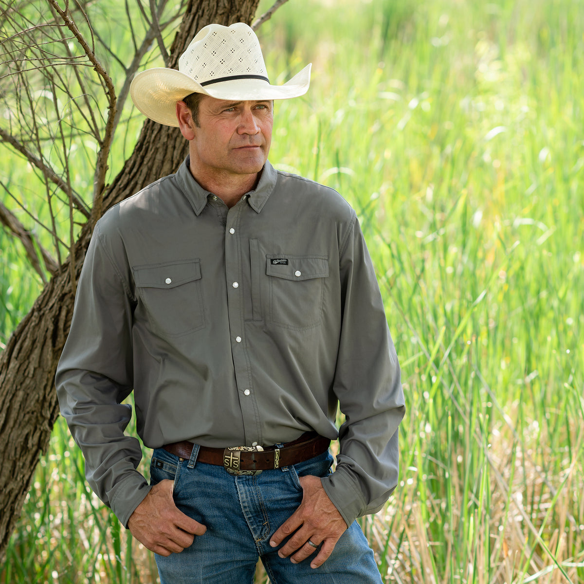 Man wearing a white cowboy hat, jeans and a long-sleeved men's shirt in a solid color, featuring a Pearl snap front closure and three exterior pockets standing in a field.