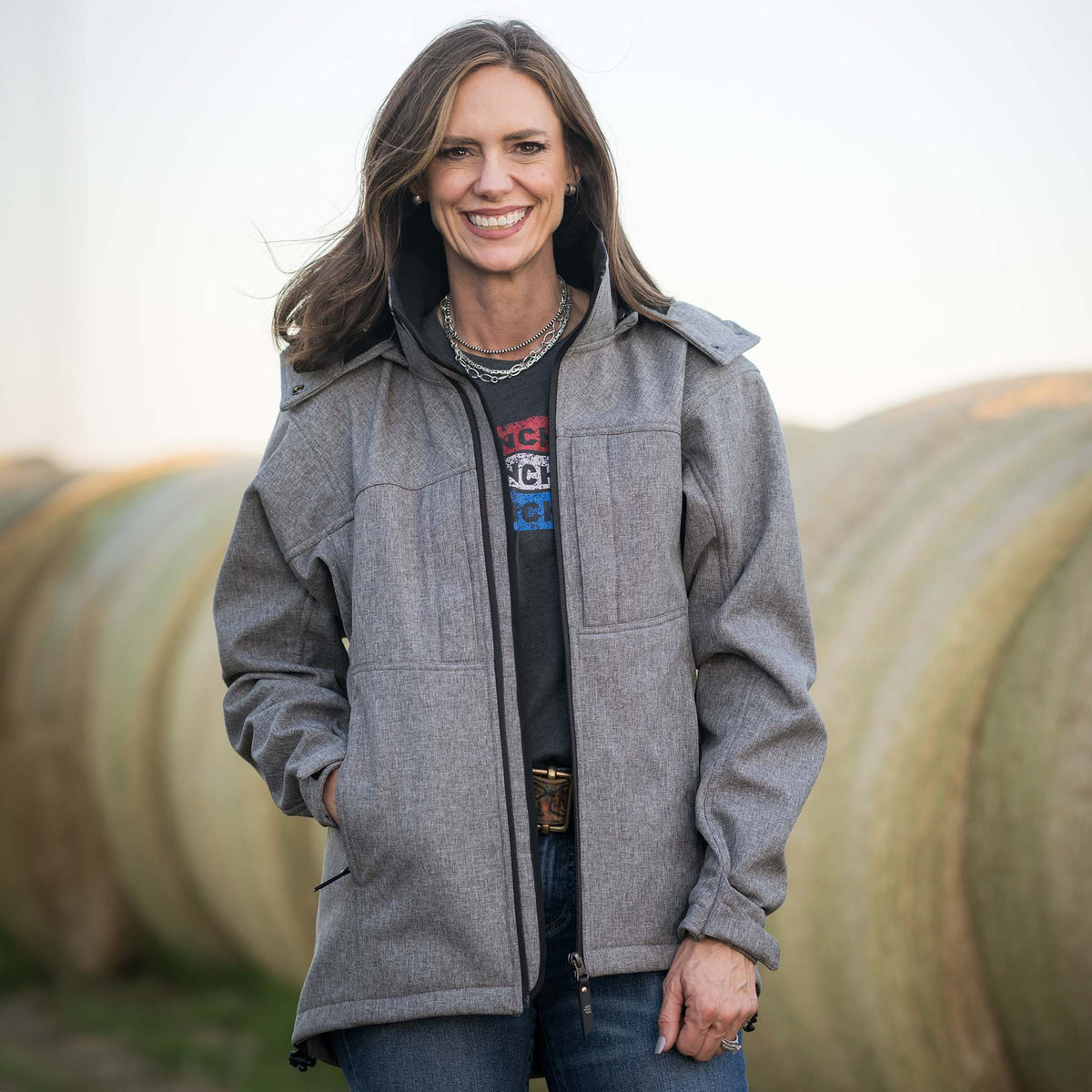Jen Smith standing in front of rolled hay bales wearing a heather gray softshell jacket with zip front closure, removable hood, and multiple pockets.