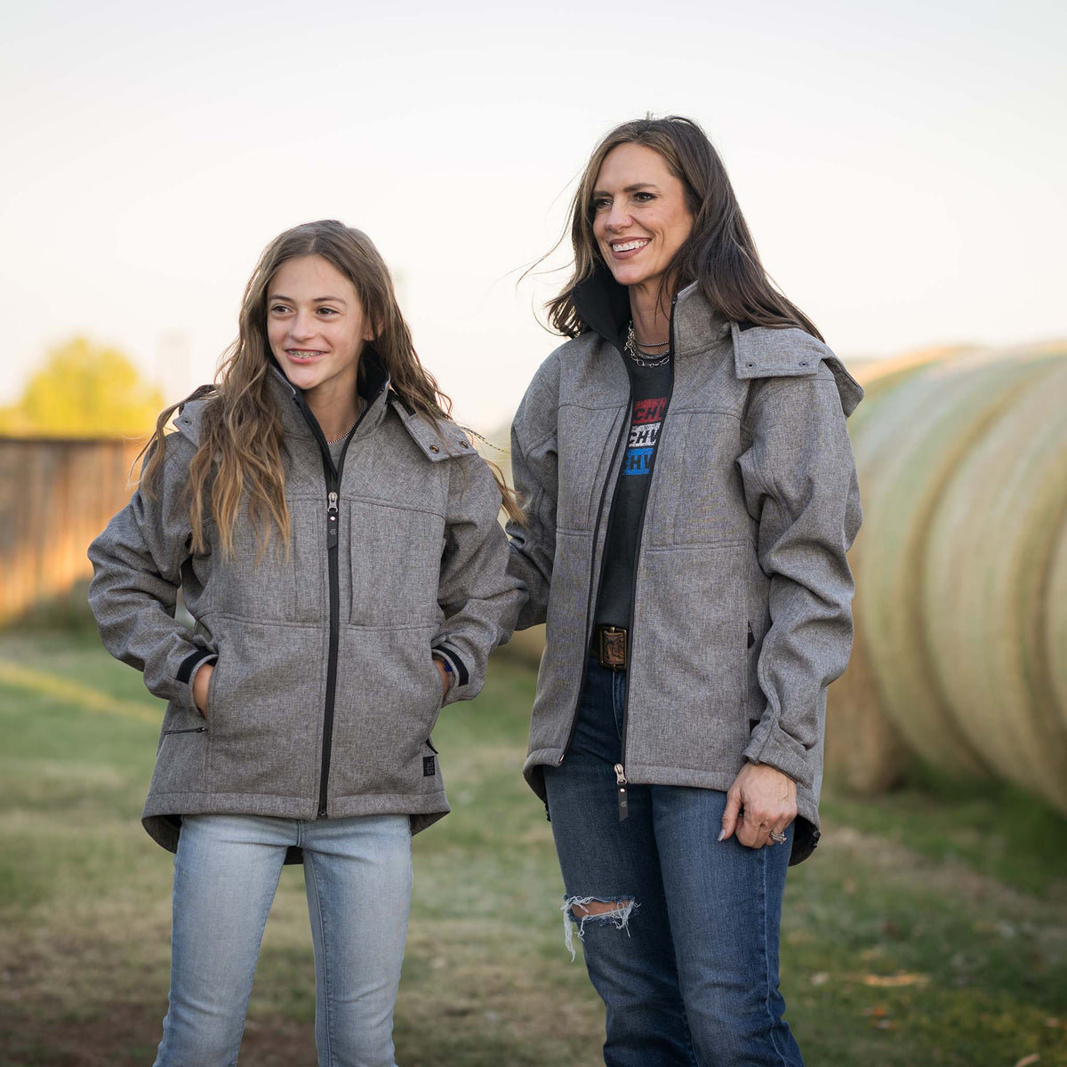 Jen and Selah Smith standing in front of rolled hay bales wearing heather gray softshell jackets with zip front closure, removable hood, and multiple pockets.