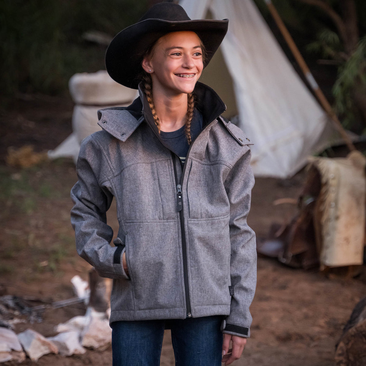 Selah Smith in a forest setting wearing a black cowboy hat and heather gray softshell jacket with zip front closure, removable hood, and multiple pockets.