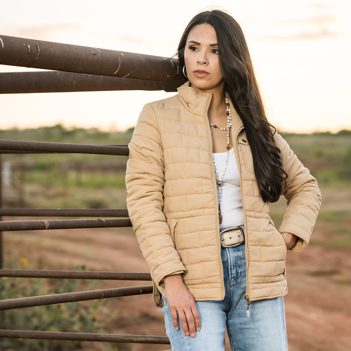 Soni Vest wearing our quilted beige jacket with zip front closure leaning against metal fence. 
