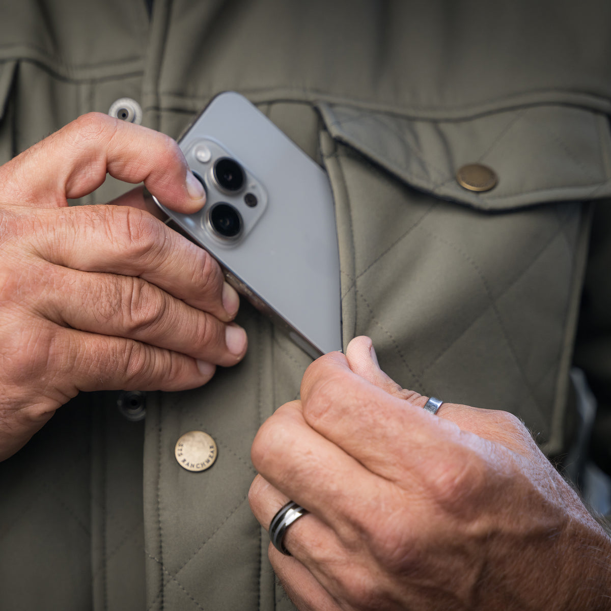 Closeup of a phone being put in the chest pocket of our olive green jacket.