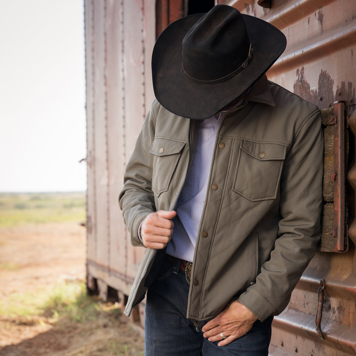 Person outside of sliding metal doors wearing our olive green jacket and black cowboy hat.