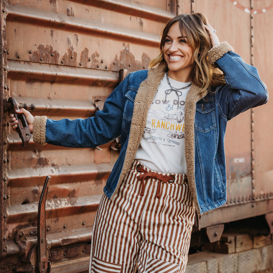 Chenae Vest wearing a light wash denim jacket lined with sherpa, two chest pockets and front button closure jacket outside of metal doors.