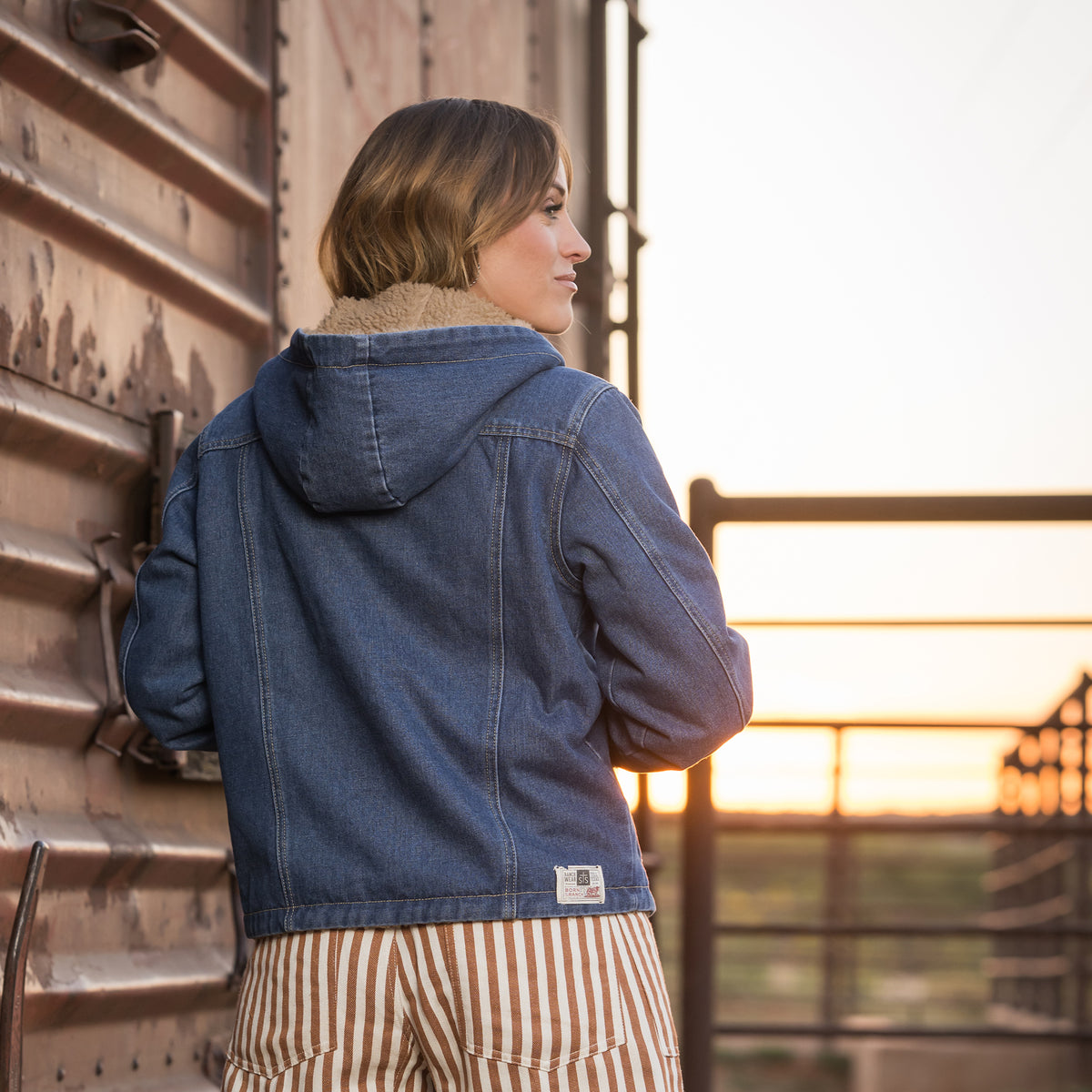 back of Chenae Vest wearing our light wash denim lined with sherpa, two chest pockets and front button closure jacket outside of metal doors