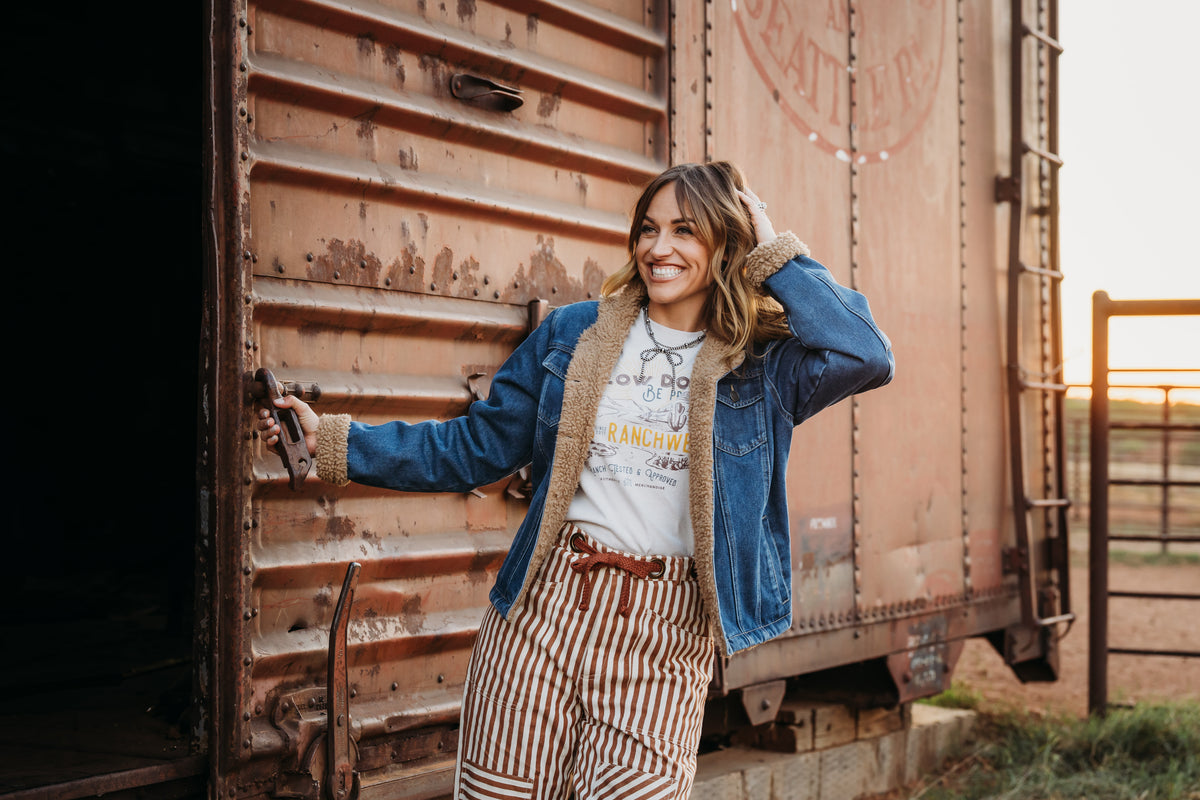 Chenae Vest wearing a light wash denim jacket lined with sherpa, two chest pockets and front button closure jacket outside of metal doors.
