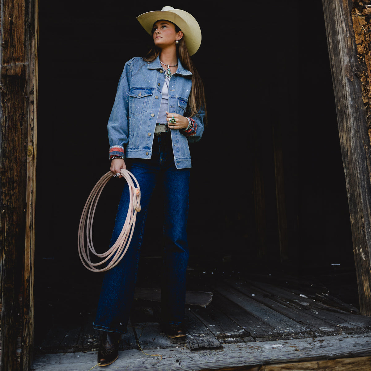 A woman wearing a light blue denim jacket, dark blue jeans, a cream cowboy hat, and holding a coiled rope stands in the doorway of a rustic wooden barn.