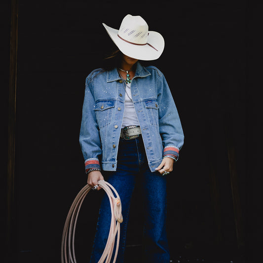 A person wearing a white cowboy hat, a light blue denim jacket stands against a dark background while holding a coiled rope.
