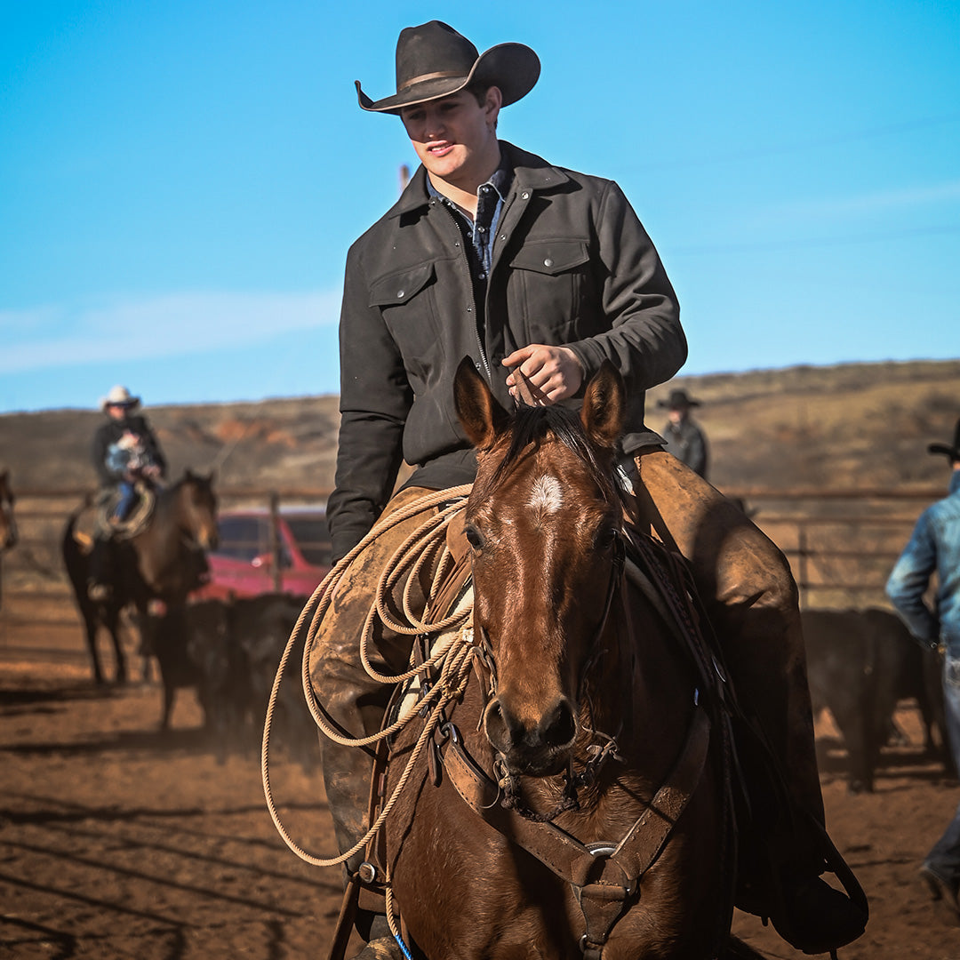 Man wearing a black cowboy hat and black softshell jacket riding a brown horse in a field in Tell, Texas.