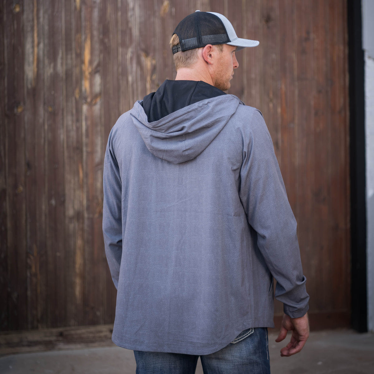 Man facing away wearing a heather gray men's rain jacket with a hood in front of a wooden building.
