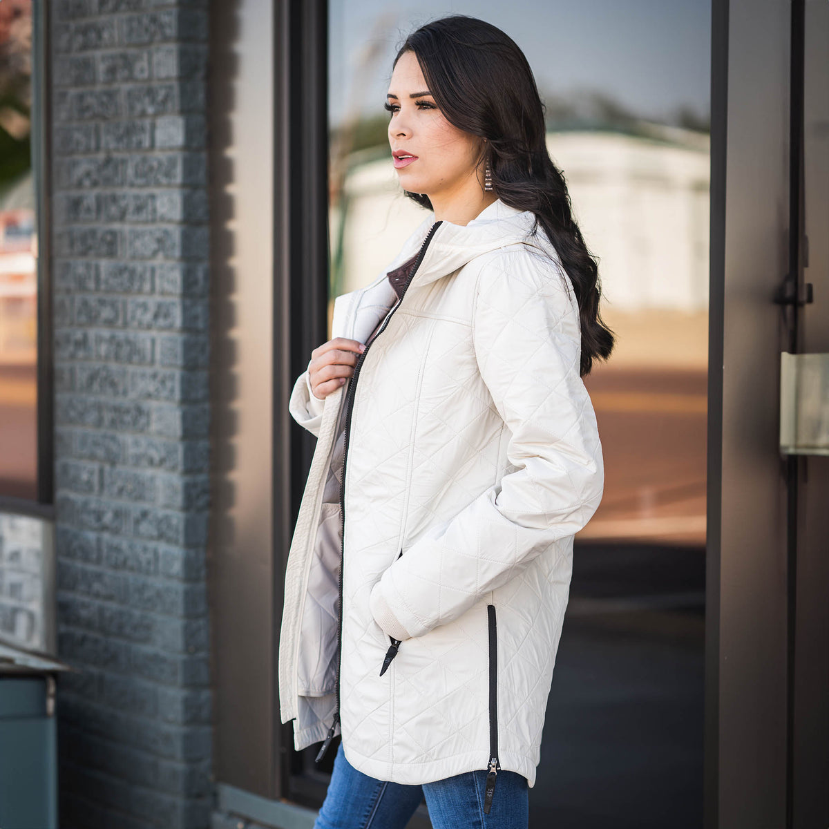 Woman wearing a beige, hooded Blakely jacket with zip front closure and ribbed cuffs standing in front of a building.