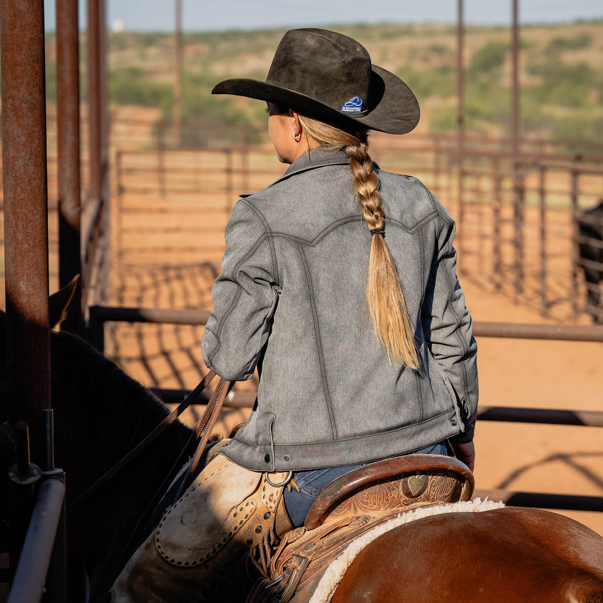 Woman facing away sitting on a horse wearing a black cowboy hat and gray softshell jacket.