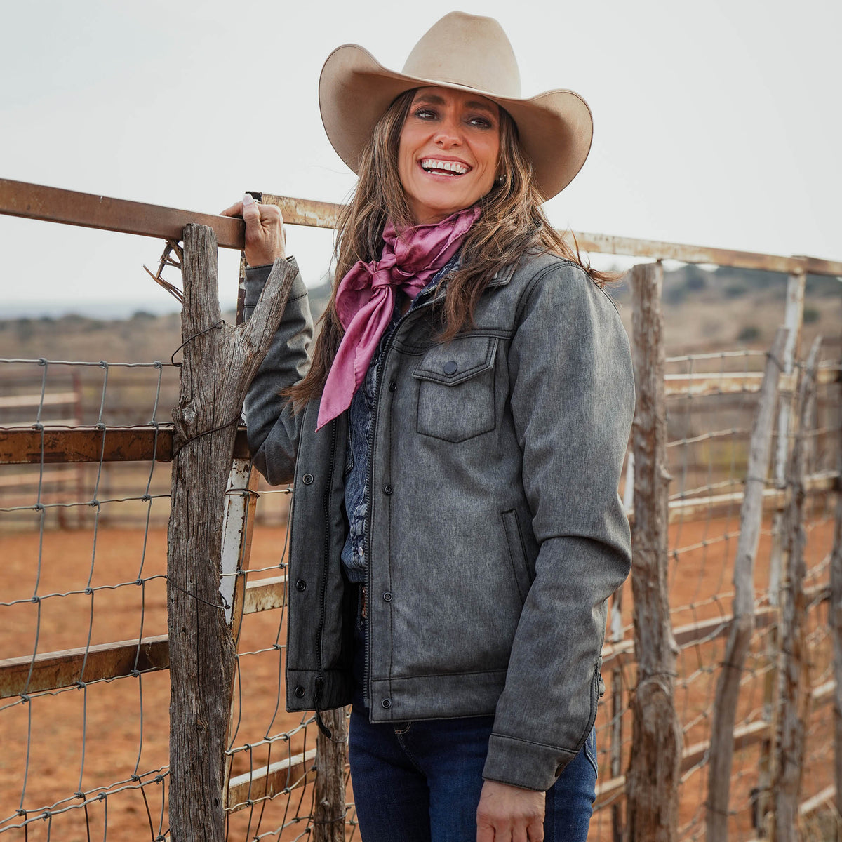 Jen Smith leaning against a fence post wearing a tan cowboy hat, jeans, pink scarf and gray softshell jacket.