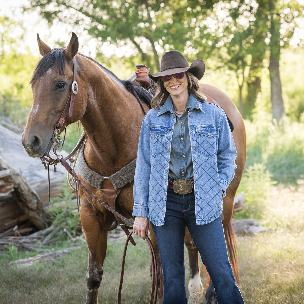 Woman in a denim jacket and cowboy hat standing next to a horse in a natural setting.