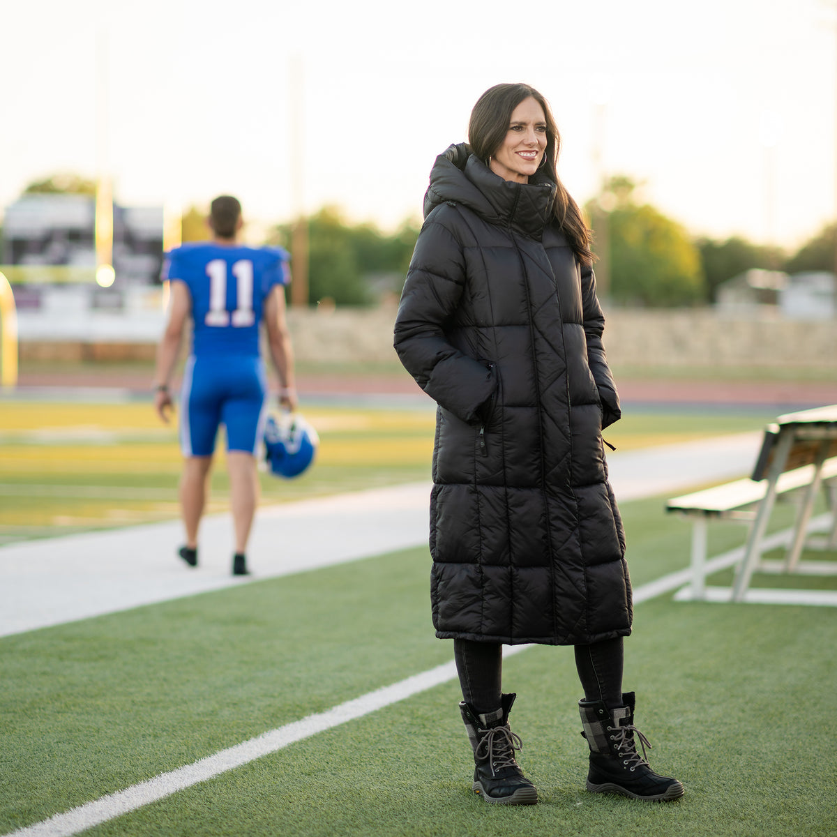 Jen Smith standing on the sidelines of a football field wearing a black puffer parka coat with zip closure hand warming pockets and hood.