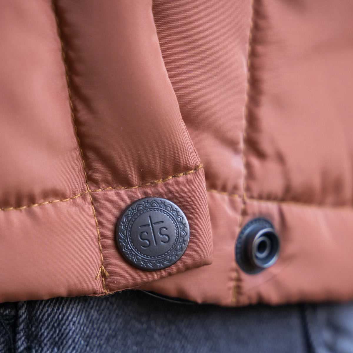 Closeup of snap on our rust collared puffer jacket with sts stamped into button.