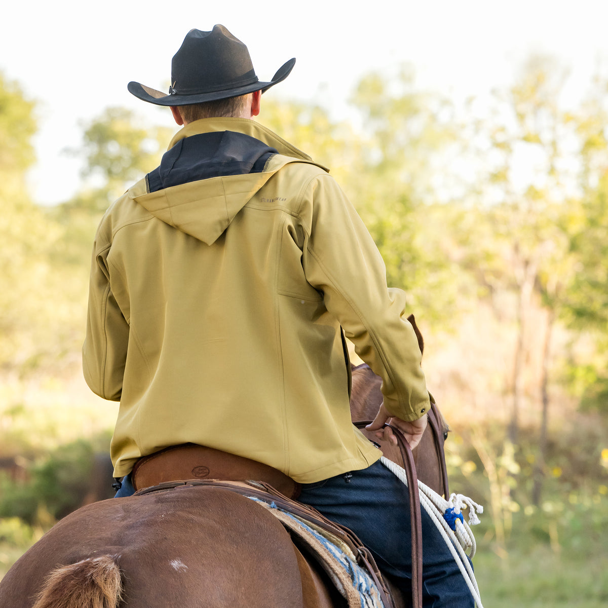 back of Clif Cooper wearing our curry yellow jacket with zip closure, removable hood and hand warming pockets outside on a brown horse