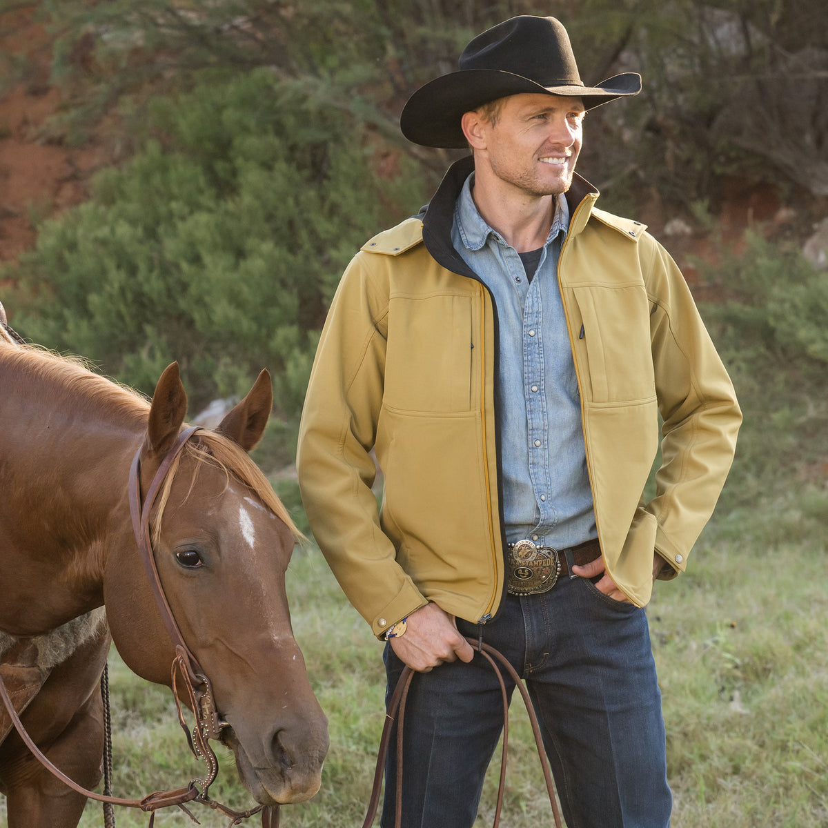Clif Cooper wearing our curry yellow jacket with zip closure, removable hood and hand warming pockets outside in front of a brown horse