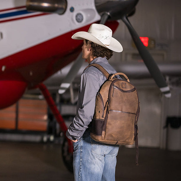 Man with a cowboy hat and backpack standing in front of a small airplane in a hangar.