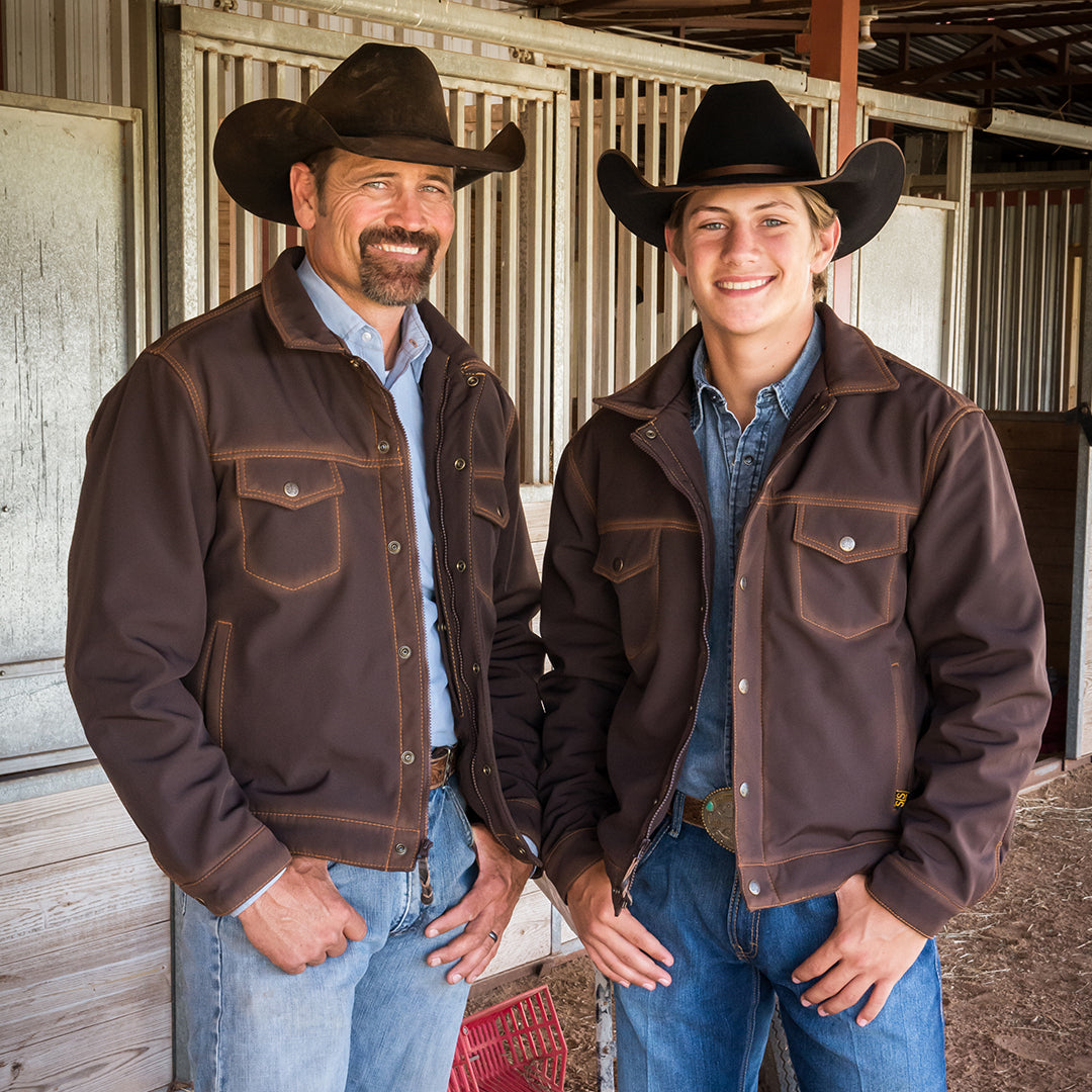 Stran T Smith and Scout Smith wearing brown softshell jackets, blue jeans and black cowboy hats standing inside a barn.