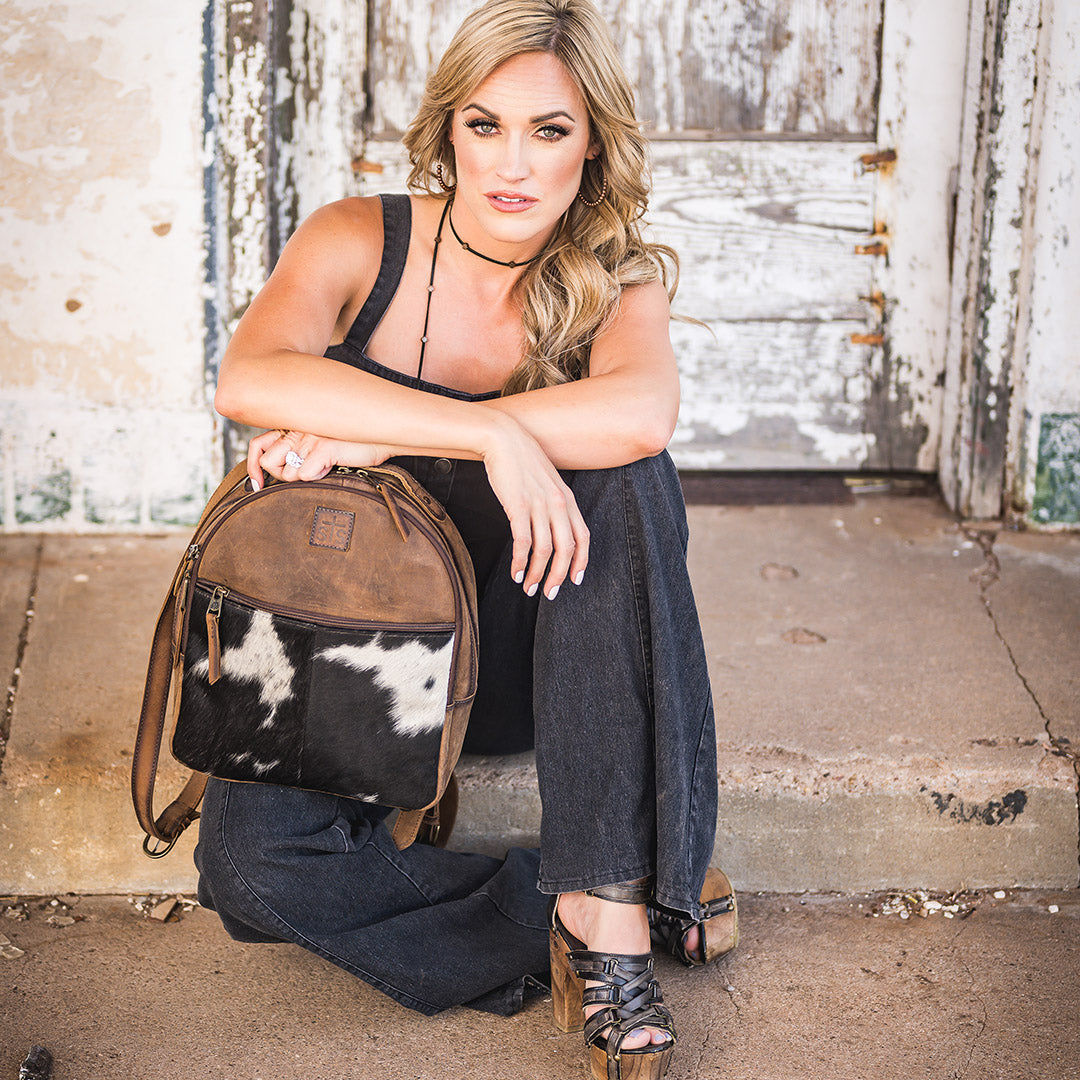 Woman wearing a black denim romper sitting on a sidewalk holding a genuine leather and hair on hide backpack with black and white hair on hide accents.