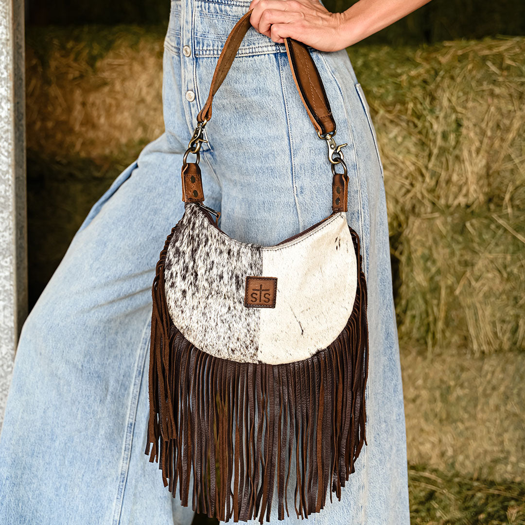 Person wearing a denim romper holding a genuine leather and hair on hide cowhide shoulder bag with fringe by the shoulder strap.