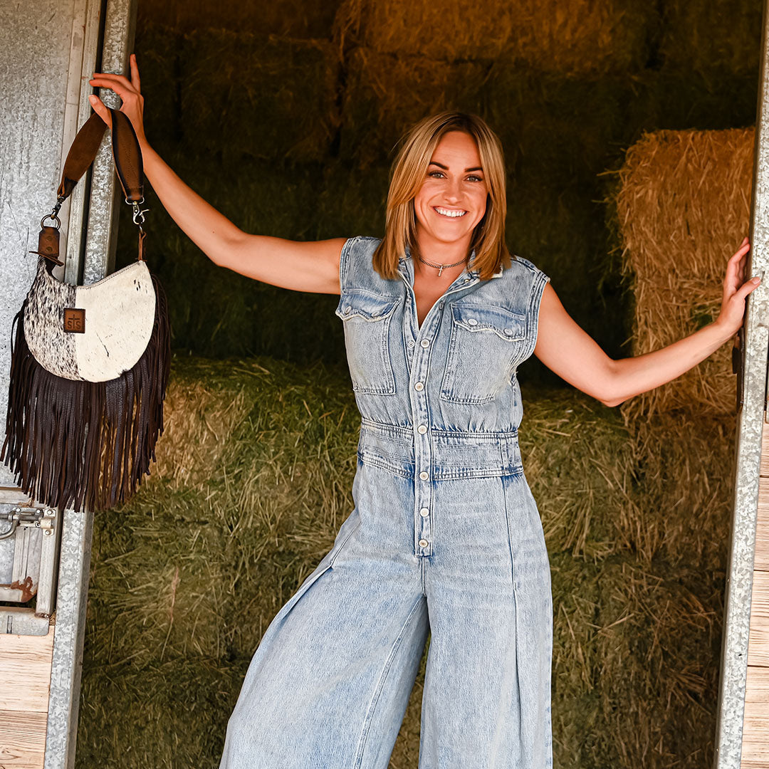 Woman wearing a denim romper leaning against a door frame holding a genuine leather and hair on hide cowhide shoulder bag with fringe.