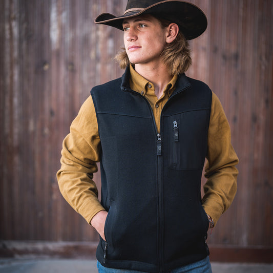 Man wearing a black cowboy hat, yellow long sleeve shirt and a black zip closure softshell vest with their hands in the vest pockets in front of a wooden wall.