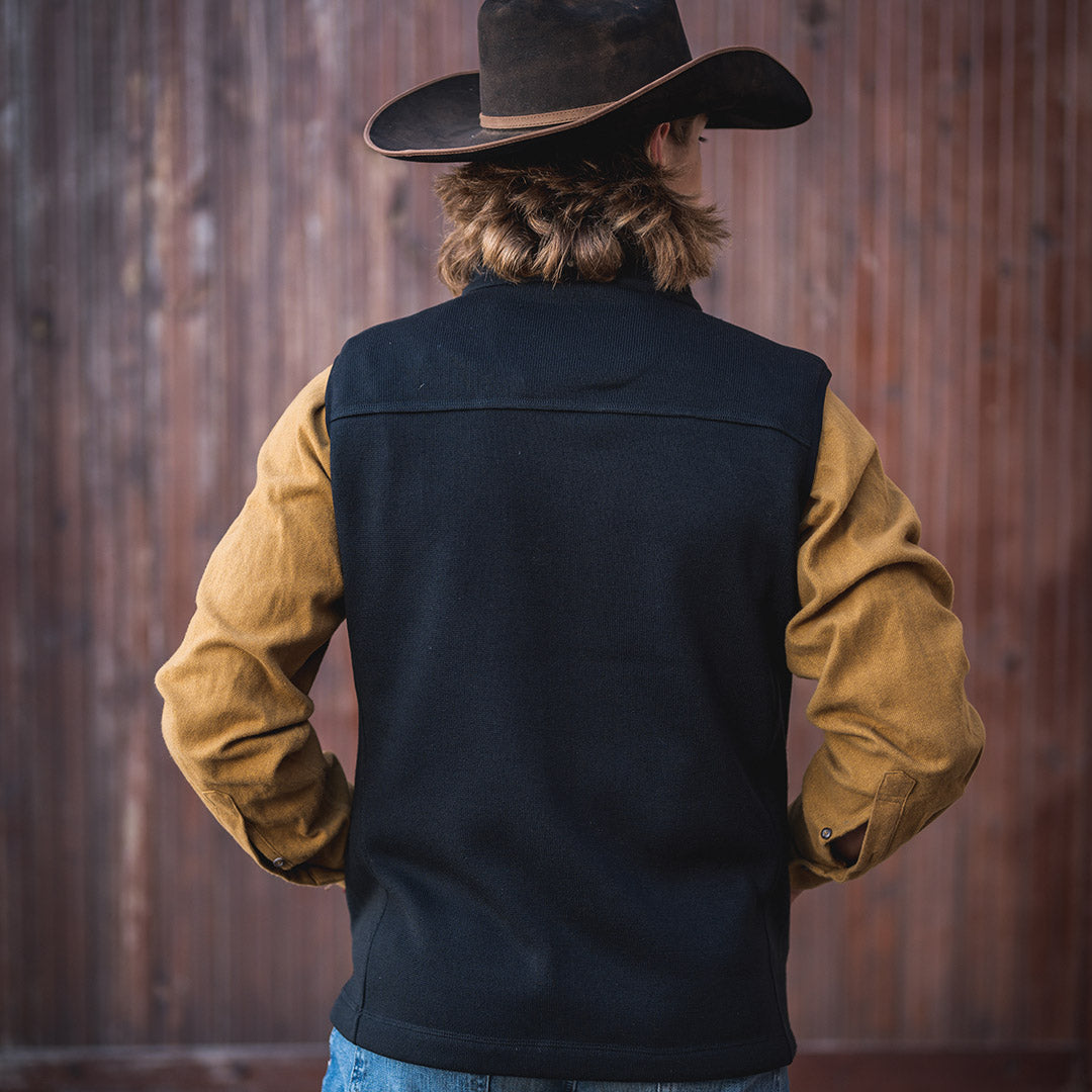 Man facing away wearing a black cowboy hat, yellow long sleeve shirt and a black softshell vest with their hands in the vest pockets in front of a wooden wall.