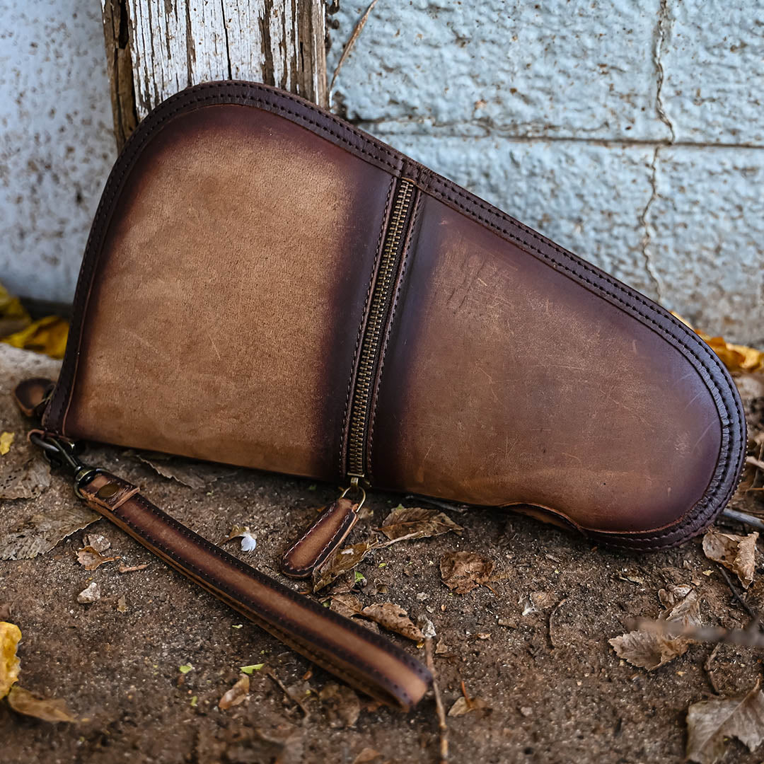Brown leather pistol case with leather wristlet strap, zip closure pocket, zip around closure sitting on dirt outside.