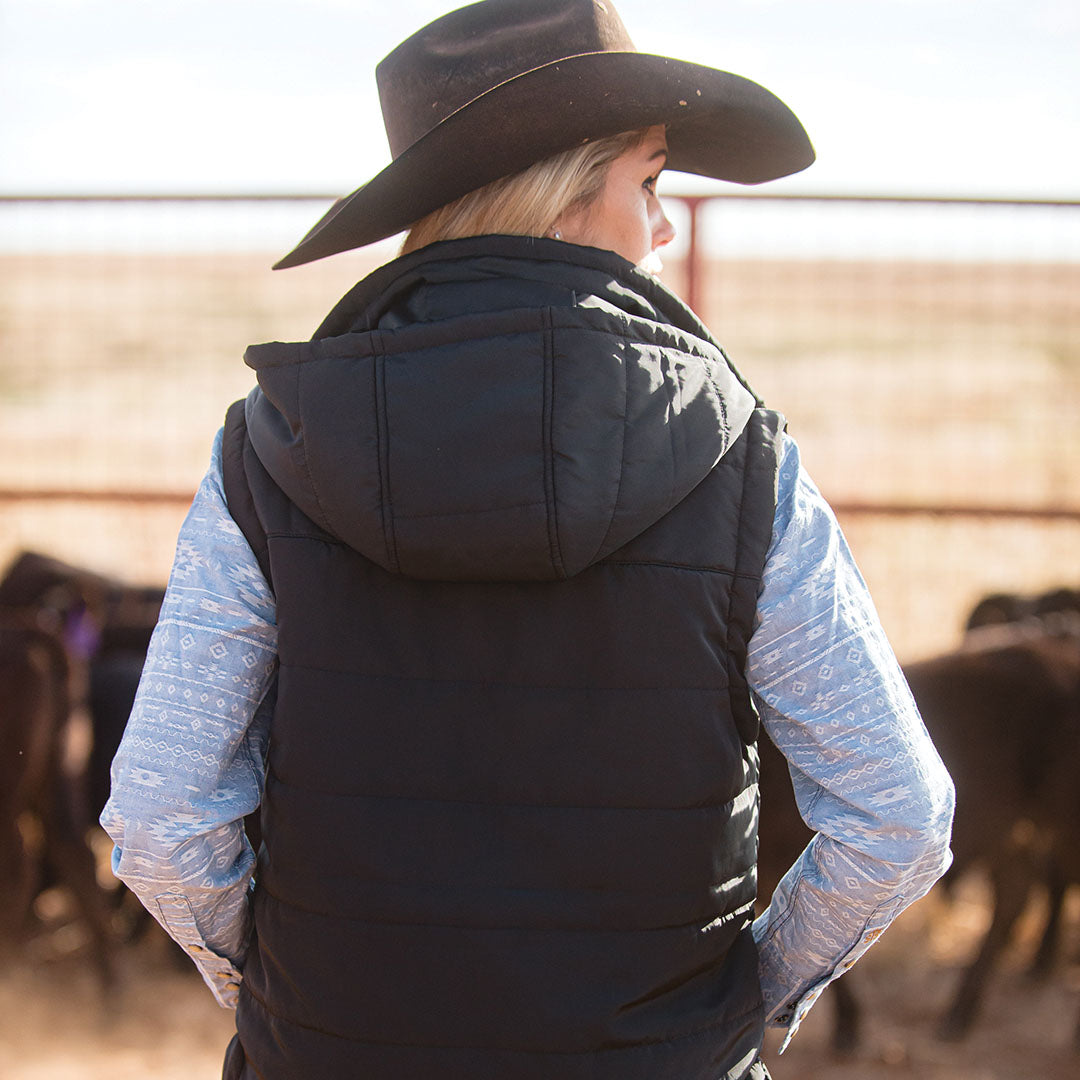Woman facing away wearing a brown cowboy hat and black puffer vest with a removable hood with cows in the background in a ranch setting.