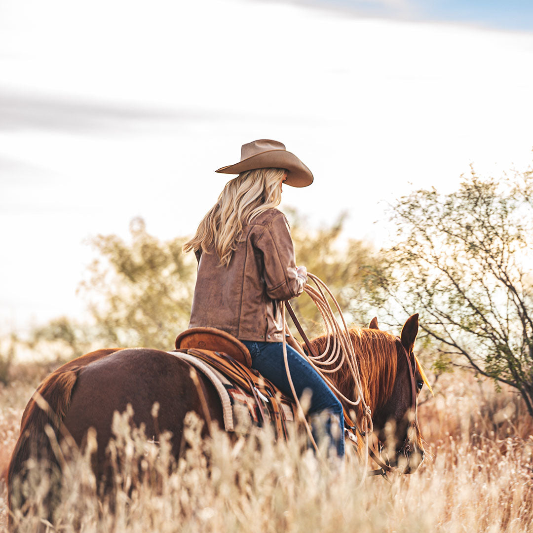 Woman wearing a tan cowboy hat, jeans and brown genuine leather jacket riding a brown horse through a field in Tell, Texas.