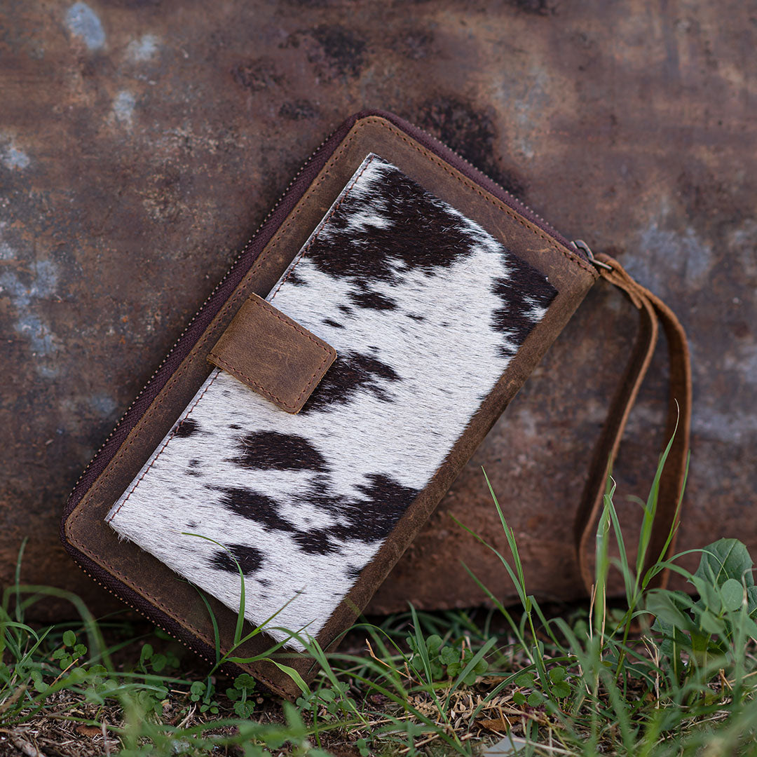 A brown cowhide Bentley wallet with genuine leather accents, zip around closure, brown leather wristlet strap and brown and white hair on hide cowhide accents on slip pocket leaning against a building in the grass.