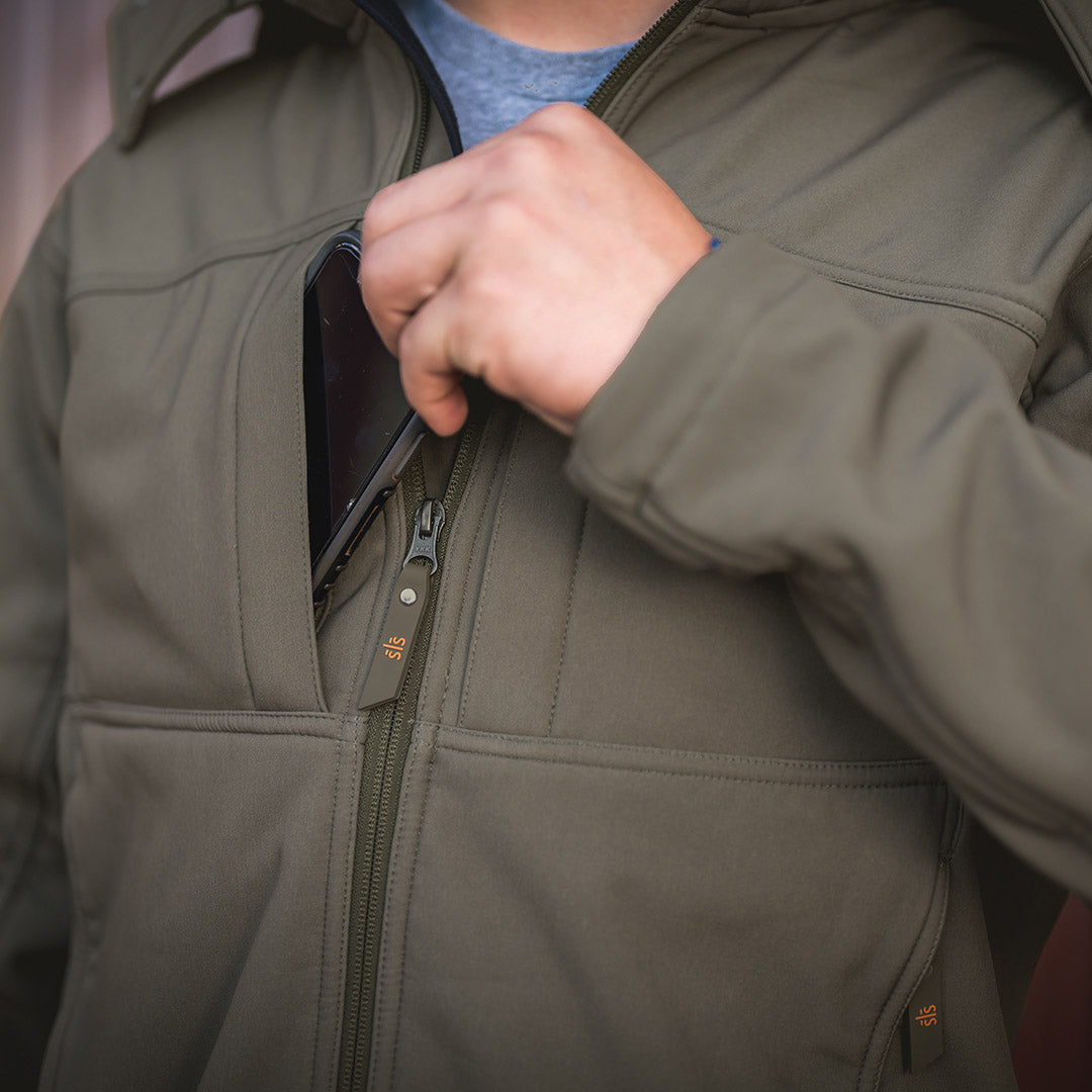 closeup of scout smith putting a cell phone in the chest pocket of our softshell olive green front zip closure jacket with removable hood in front of wooden door