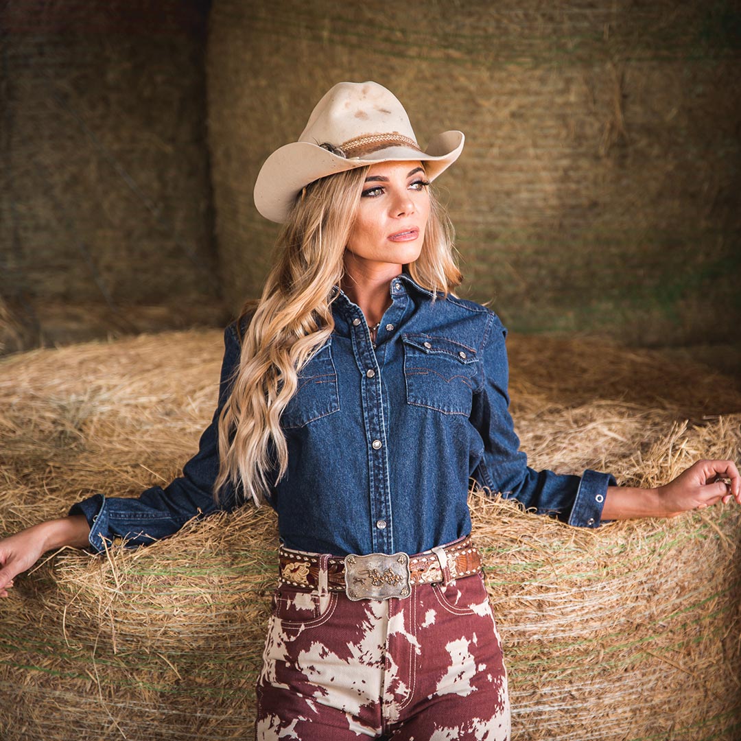Woman wearing a tan cowboy hat and classic denim pearl snap shirt leaning against a hay bale.