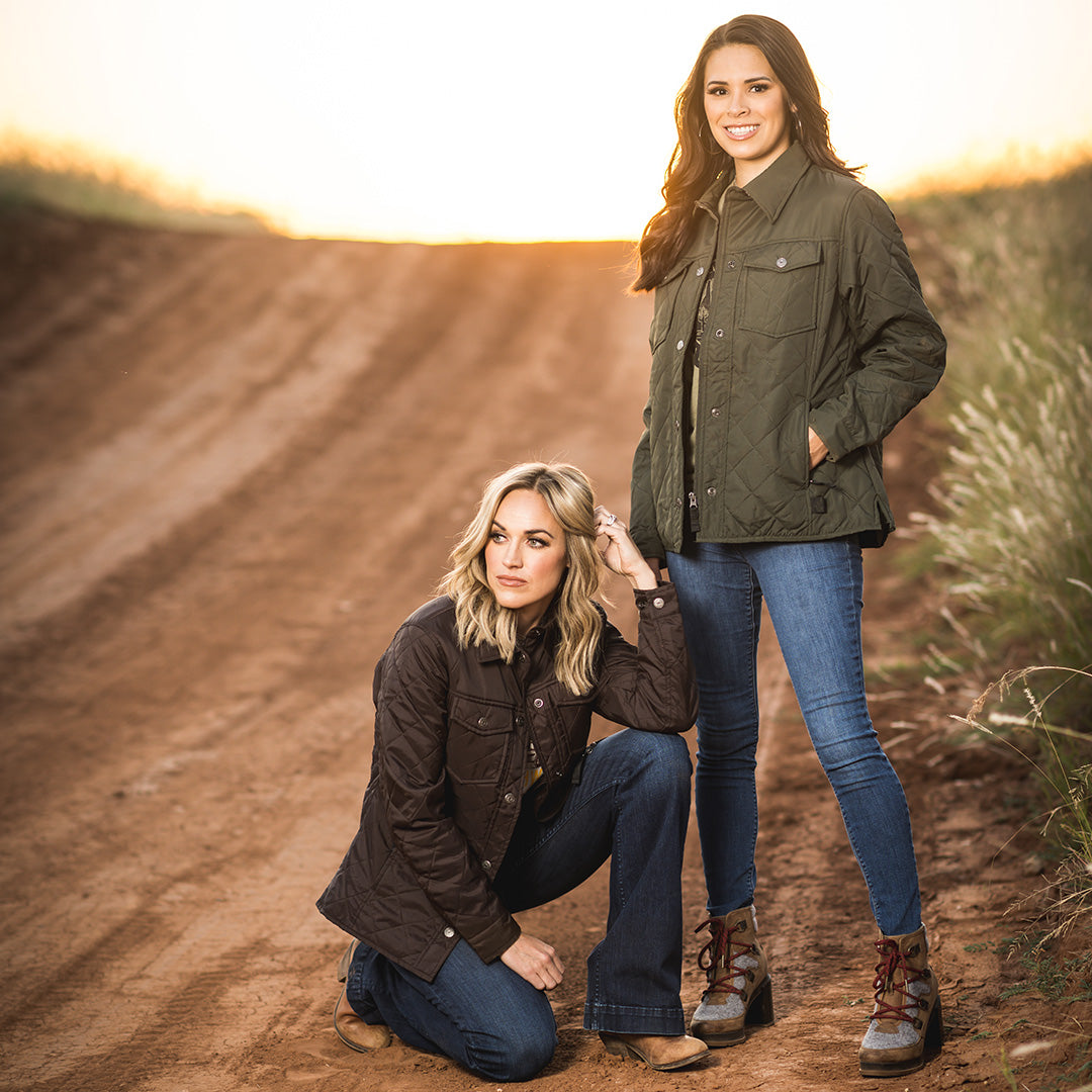 Two women wearing olive and brown softshell jackets standing on a dirt road.