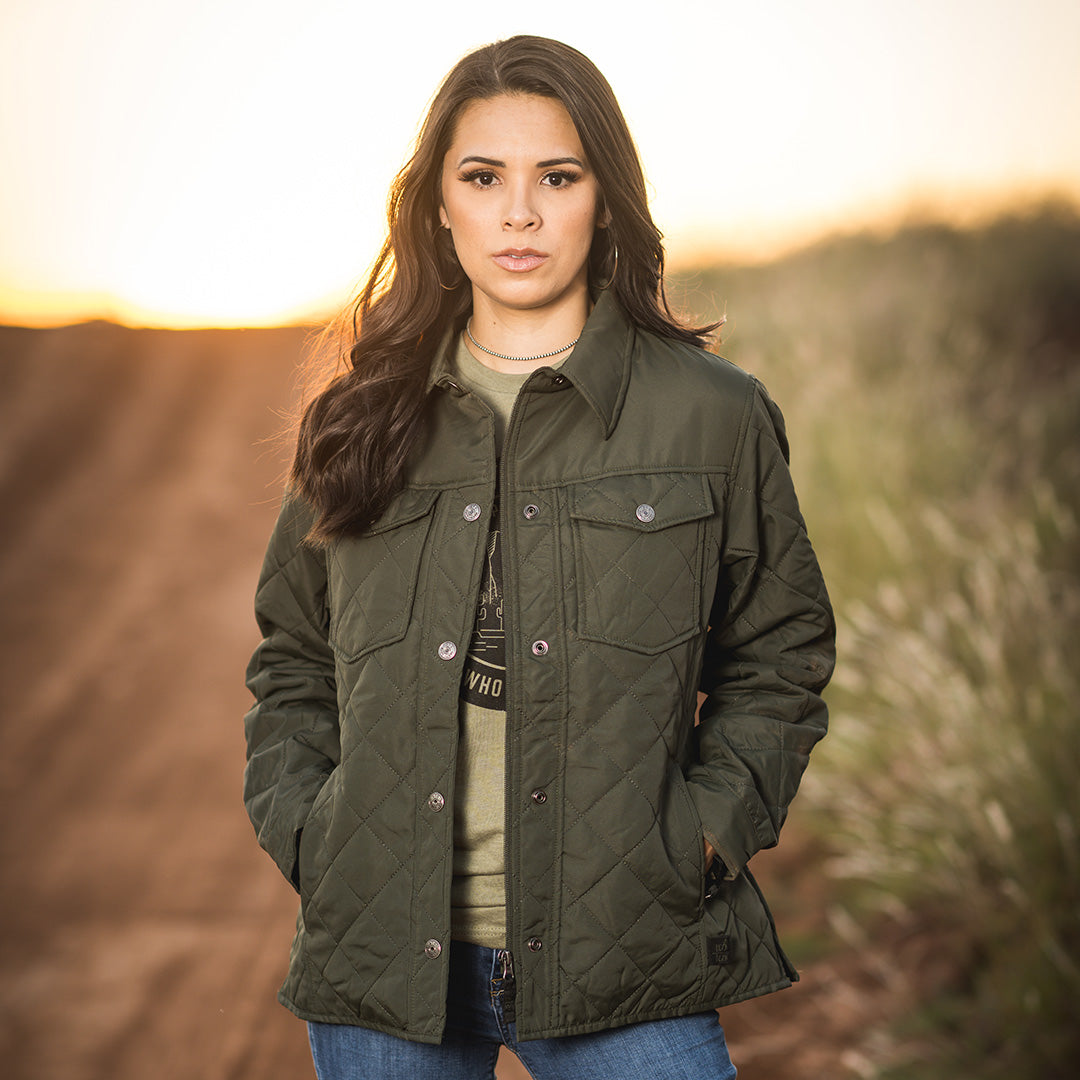 A woman wearing an olive softshell jacket standing in a field on a ranch in Tell, Texas.