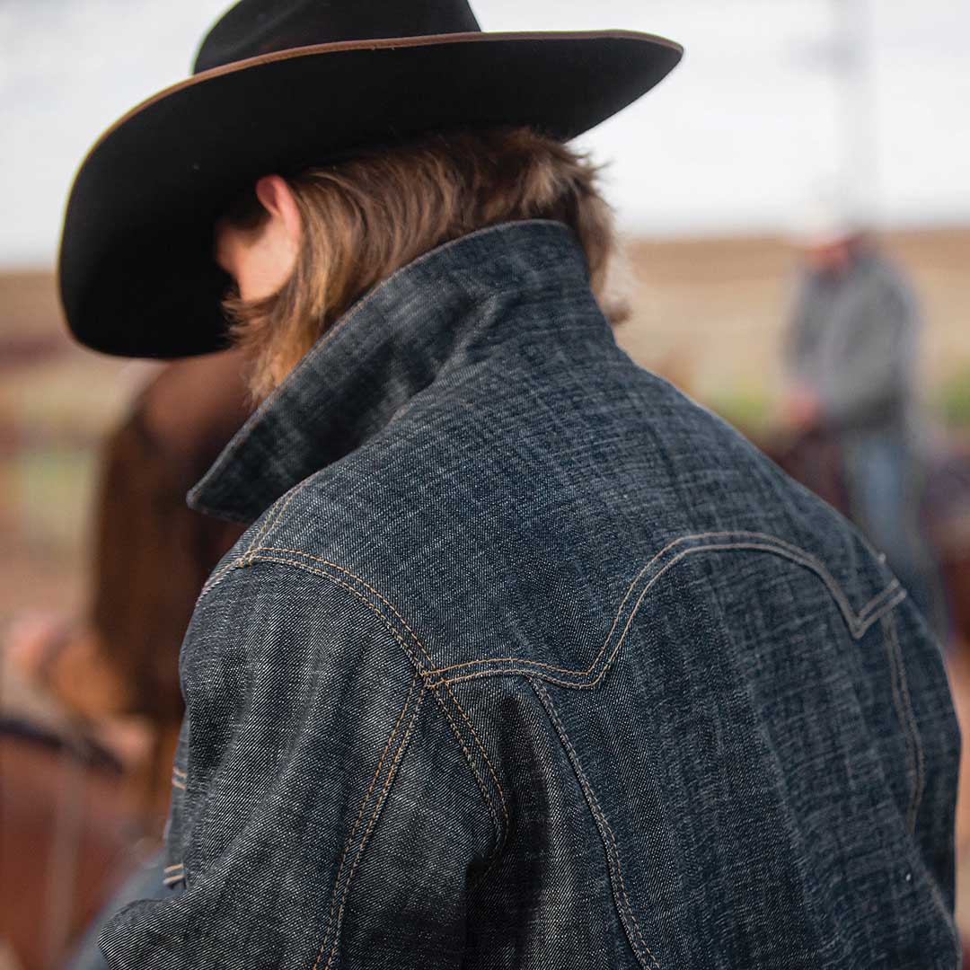Man standing in a field wearing a black cowboy hat and denim jacket.