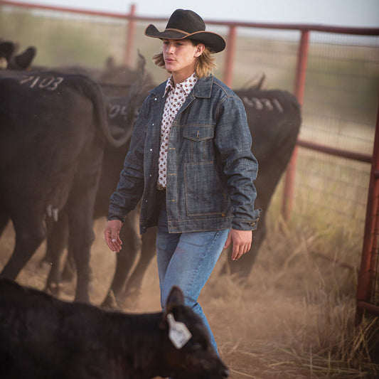Man standing in a cow pasture with cows around him wearing a black cowboy hat, jeans and a denim jacket.